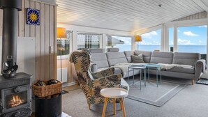 A cozy cabin interior with a wood-burning stove, a sofa, a patterned armchair, and a view of the ocean. - "Nedelko" - 20m from the sea by Interhome (Otterup)