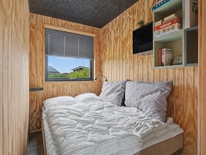 Ein Schlafzimmer mit Bett, Fenster, Holzwänden und einem Regal mit Büchern und einer Pflanze.