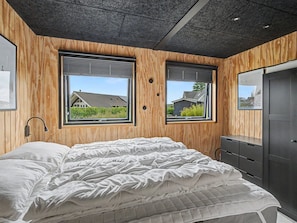 Ein Schlafzimmer mit Bett, Holzwänden, zwei Fenstern und einem Kleiderschrank.