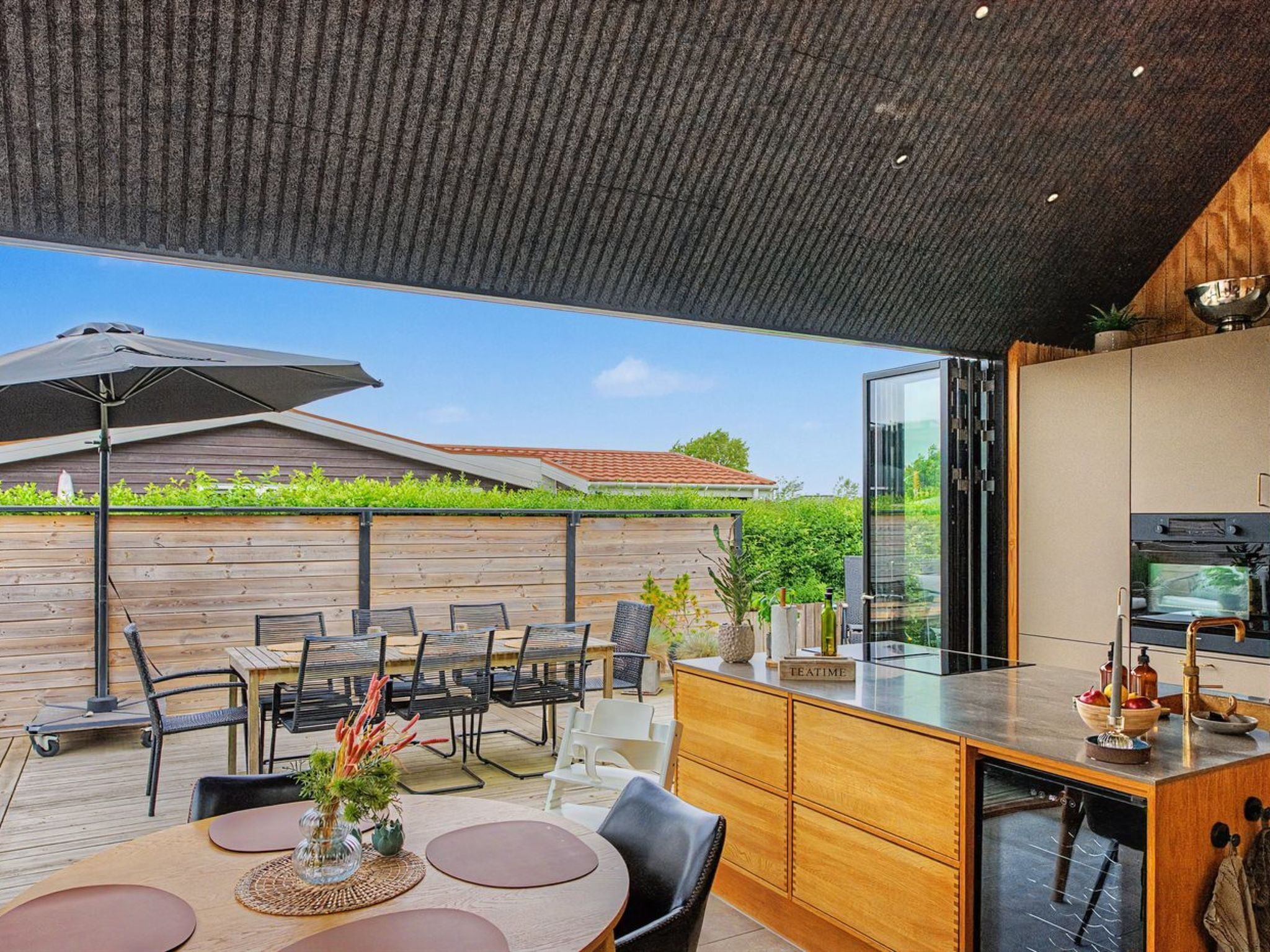 A modern outdoor kitchen with a dining area, including a table with chairs, a wooden deck, and a view of a house in the background.