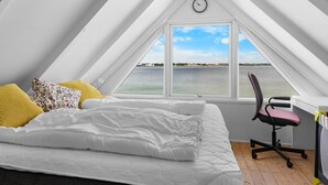Ein Schlafzimmer mit einem großen Bett, einem Schreibtisch und einem Stuhl, mit Blick auf ein Gewässer durch ein Fenster.
