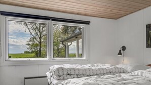 Un dormitorio con una cama, un radiador, una ventana con vistas a árboles y un campo, y una lámpara fijada a la pared.