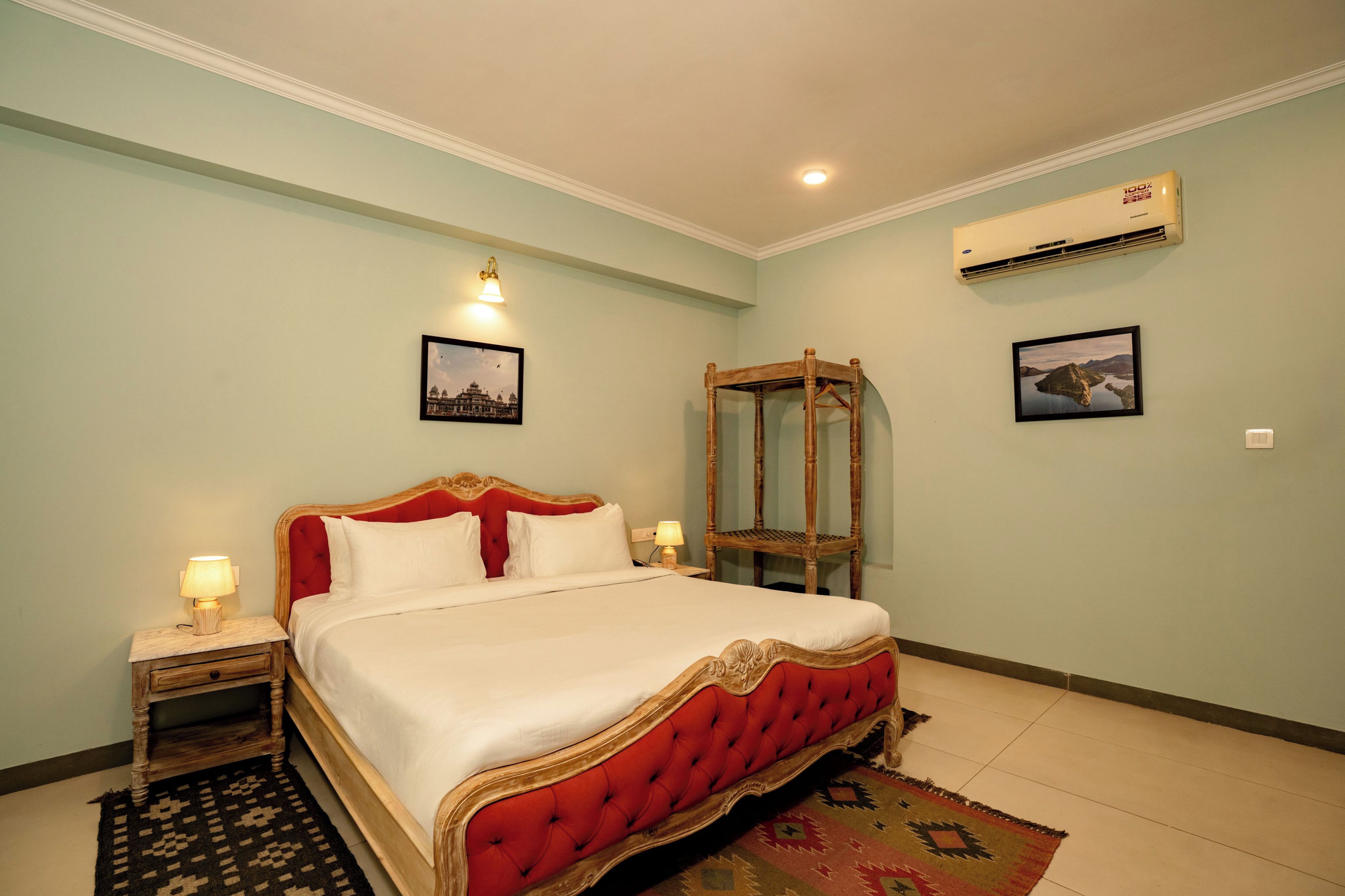 A hotel room with a large bed, a nightstand, a bedside lamp, a wall-mounted air conditioner, and two framed pictures.