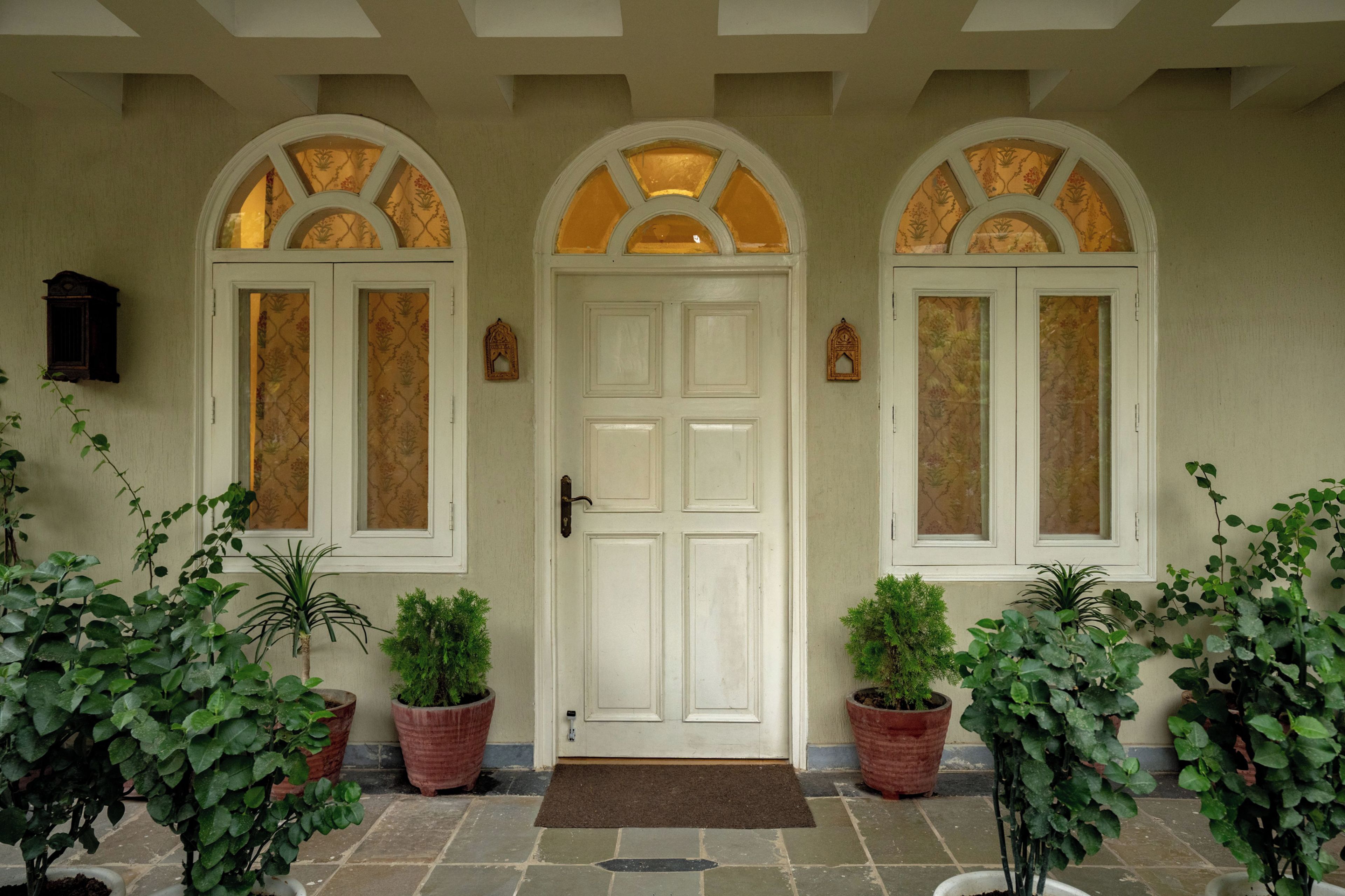 Puerta blanca con felpudo marrón, flanqueada por dos macetas y dos ventanas con vidrieras decorativas.