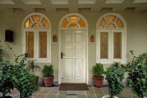 Puerta blanca con felpudo marrón, flanqueada por dos macetas y dos ventanas con vidrieras decorativas.