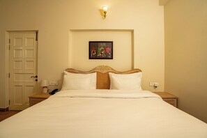 A hotel room with a bed, a nightstand, a lamp, a painting, and a door. - Ramat Niwas by Kutani Stays (Jaipur)