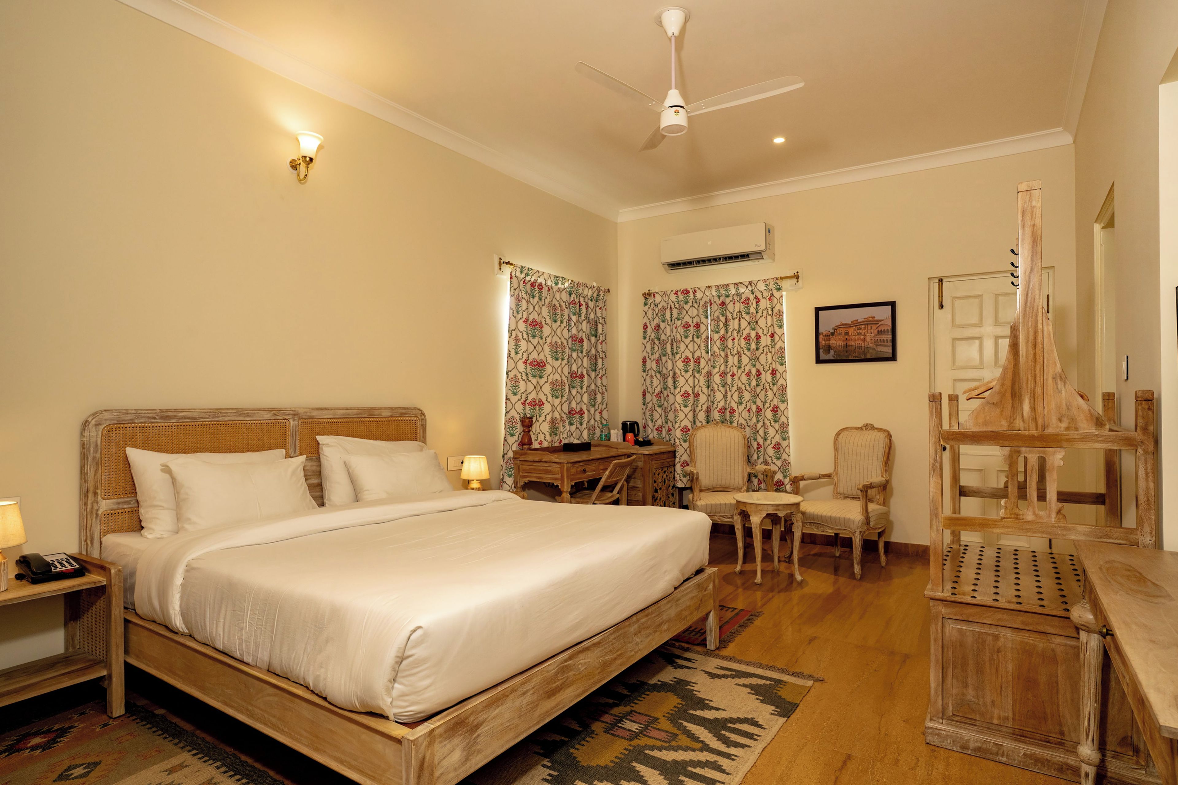A hotel room with a wooden bed, a desk, two chairs, a small table, a ceiling fan, and a wall-mounted air conditioner.