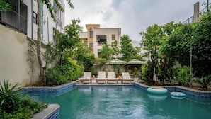 Una piscina con sillones de descanso y flotadores, rodeada de vegetación y edificios.