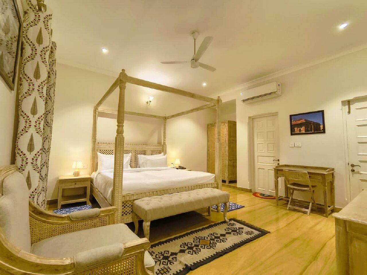 A four-poster bed with a canopy, a wooden desk, a chair, and a small bedside table with a lamp.