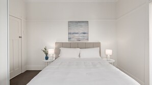 Ein Hotelzimmer mit einem großen Bett, zwei Nachttischen mit Lampen, einem Bild an der Wand und einer Tür links.