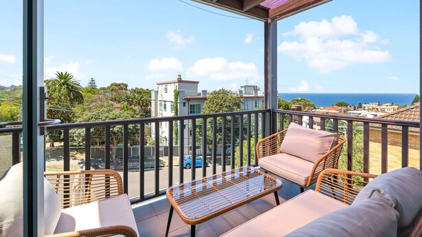 Ein Balkon mit Korbmöbeln, einem Glastisch und Blick auf das Meer und die Gebäude.