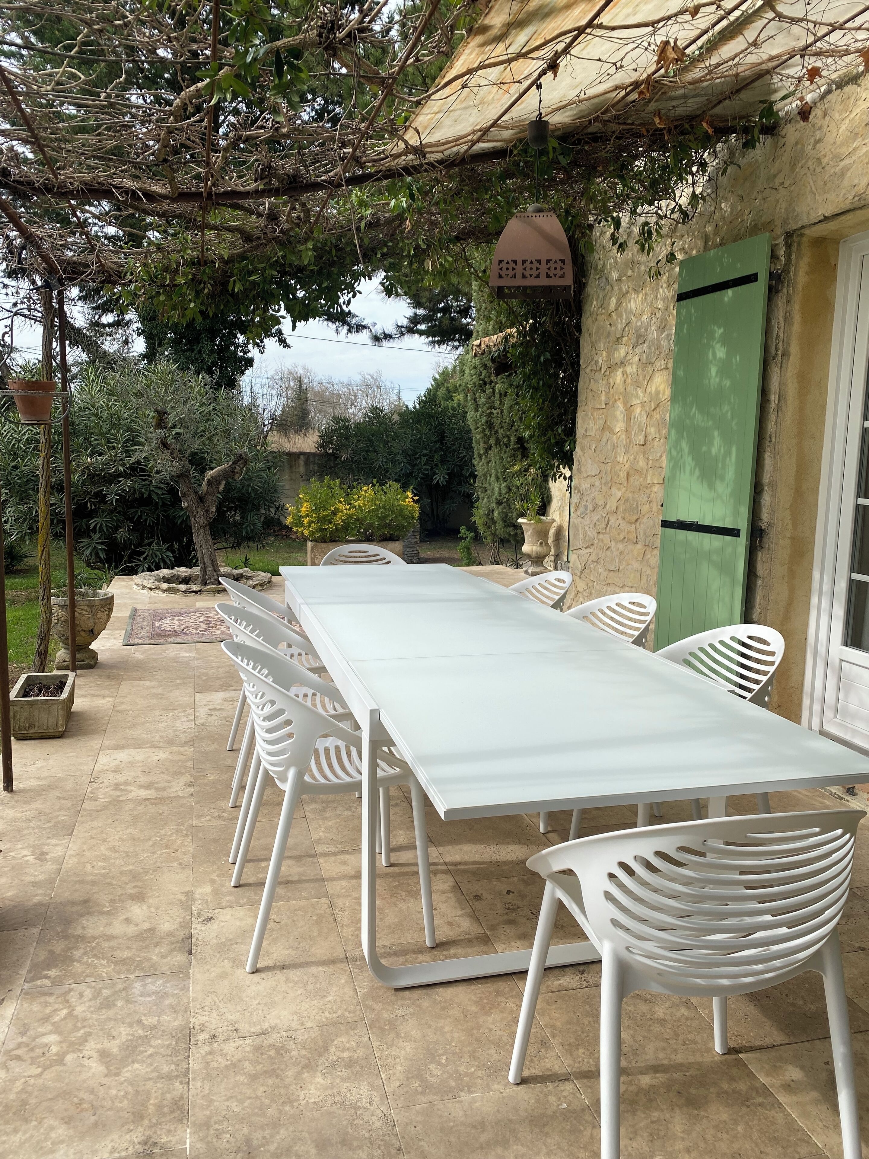 Un espace extérieur abrité, aménagé pour les repas, avec une longue table et des chaises blanches, entouré de verdure et d’un mur en pierre.