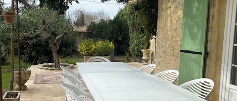 Un espace extérieur abrité, aménagé pour les repas, avec une longue table et des chaises blanches, entouré de verdure et d’un mur en pierre.