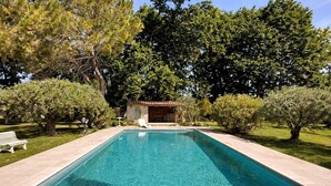 Une piscine rectangulaire à l’eau d’un bleu limpide, entourée d’une terrasse carrelée. Un banc blanc se trouve sur le côté gauche, et un petit bâtiment avec un toit en tuiles est visible au fond.