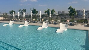 Una piscina al terrat amb aigua cristal·lina blava, envoltada de gandules i ombrelles, amb un skyline urbà al fons.