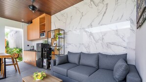 Una sala de estar moderna con un sofá gris, una mesa de centro de madera y una zona de cocina con microondas y refrigerador.