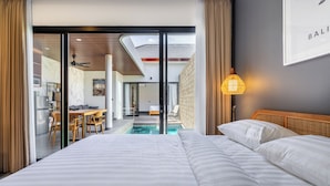 Habitación de hotel moderna con una cama grande, vistas a una piscina y un área de comedor visible a través de una puerta abierta.