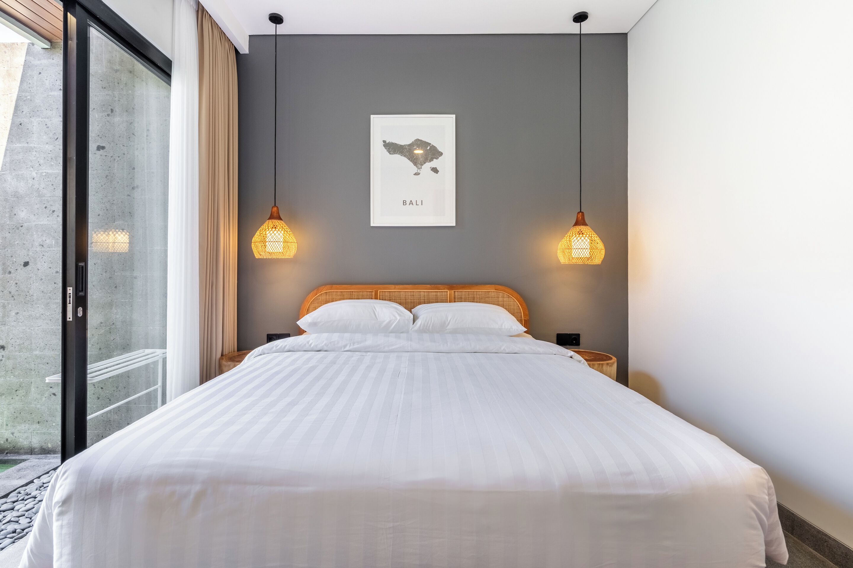 A neatly made bed with white linens, a framed map of Bali on the wall, and two hanging lamps.