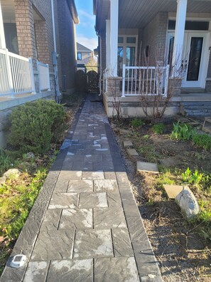 Un chemin pavé mène à une maison dont la façade est en pierre, ornée d’une balustrade blanche et bordée d’un petit jardin.