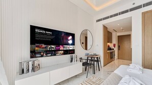 A modern hotel room with a large flat-screen TV on the wall, a round mirror, a dining area with a table and chairs, a neatly made bed, and a walk-in closet. - Elegant Studio in Azizi Riviera | Perfect Getaway (Dubai)