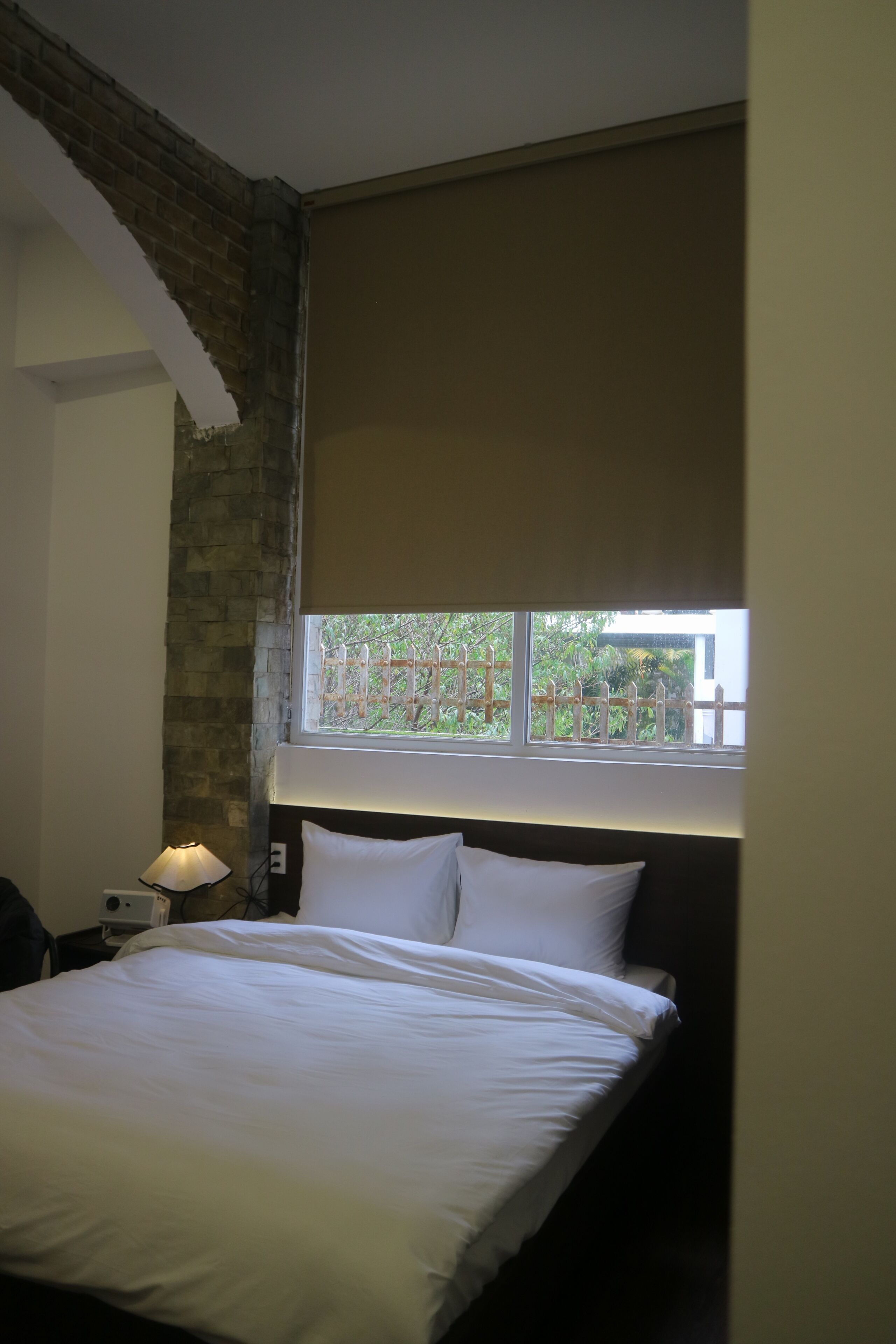 A bedroom with a bed, a nightstand, a lamp, a window with a view of greenery, and a brick wall.