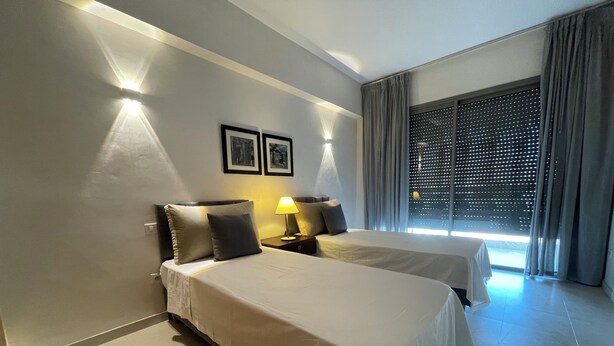 A hotel room with two beds, a nightstand with a lamp, a large window with curtains, and framed pictures on the wall. - Beautiful apartment in downtown Agadir at Galerie Talborjt (Agadir)