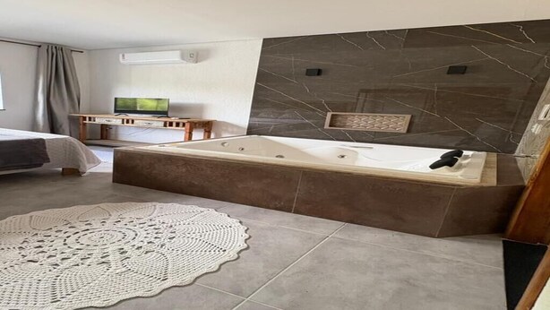 A modern bathroom with a large bathtub, a vanity with a mirror, and a television on a wooden stand. - Village House in the Valley (Arraial d'Ajuda)