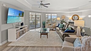 A spacious living room with a large flat-screen TV, a ceiling fan, and a view of a balcony with a cityscape. - Royale Palms 804 2 Br Oceanview in Kingston, Pool (Myrtle Beach)