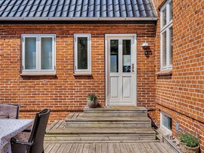 A brick house with a white door, two windows with shutters, a small porch with steps, and a table with chairs.