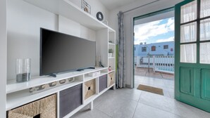 Ett modernt vardagsrum med en platt-TV, vita hyllor, korgar av rotting och utsikt över en pool och byggnader genom en grön dörr.
