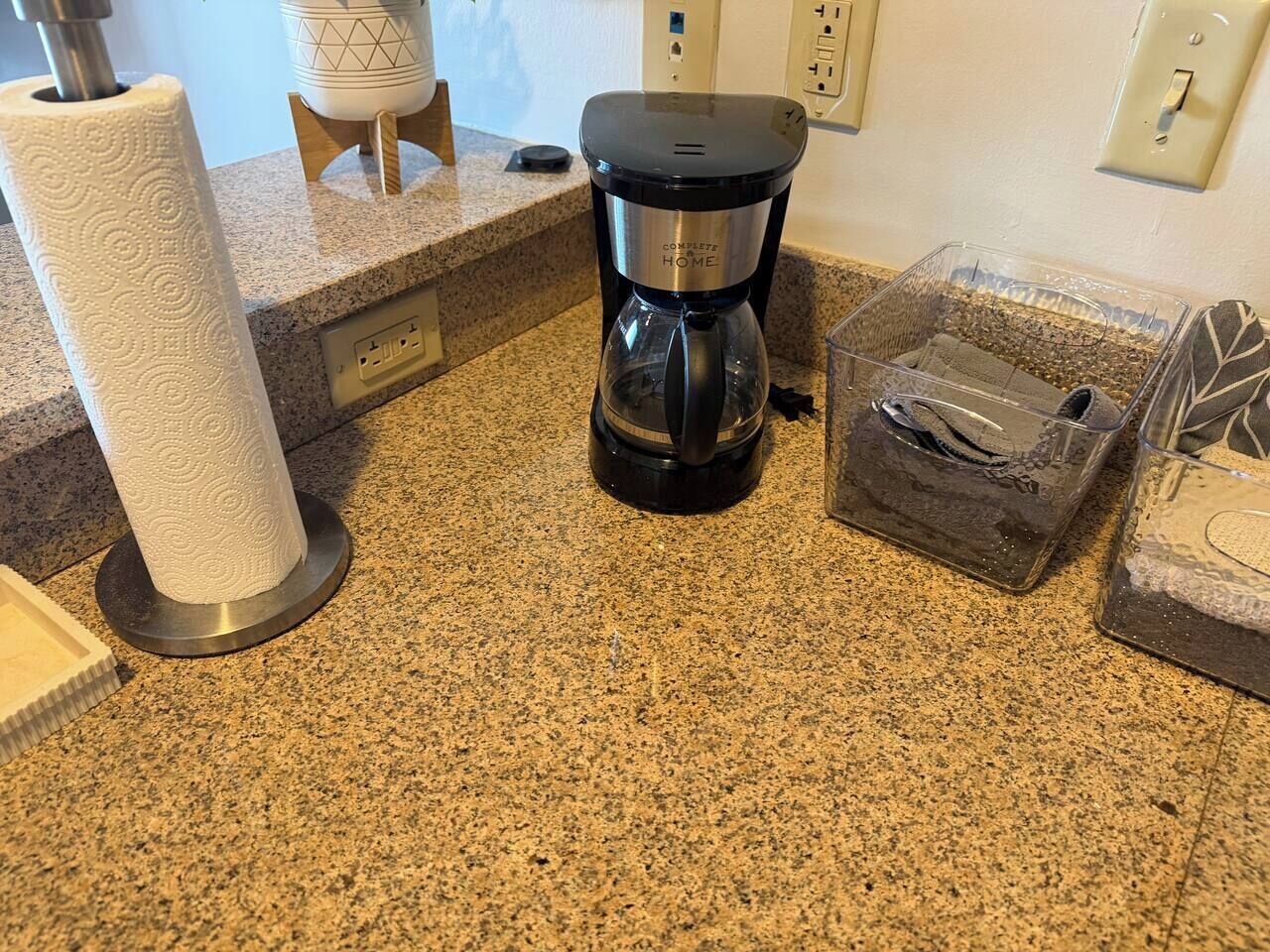 Una encimera de cocina con una cafetera, un rollo de toallas de papel y un recipiente de plástico transparente con una cuchara.