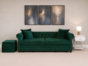 Un sofá verde de terciopelo con un otoman a juego y una mesita de noche con una lámpara, frente a una pared lisa decorada con arte abstracto.