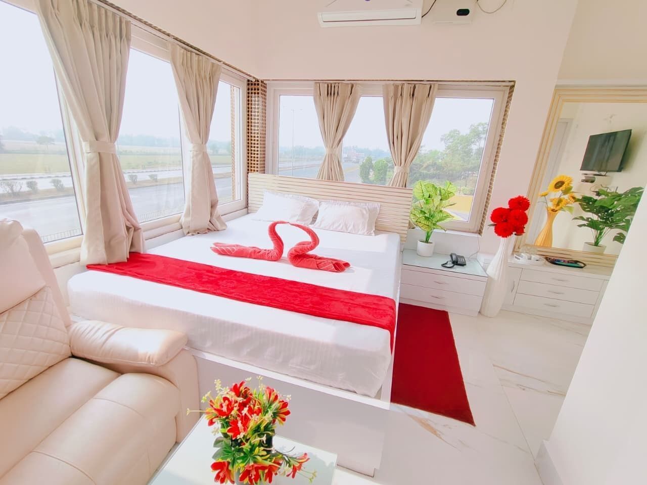 A hotel room with a bed, a sofa, a small table with a vase of flowers, and a view of the outdoors through large windows.