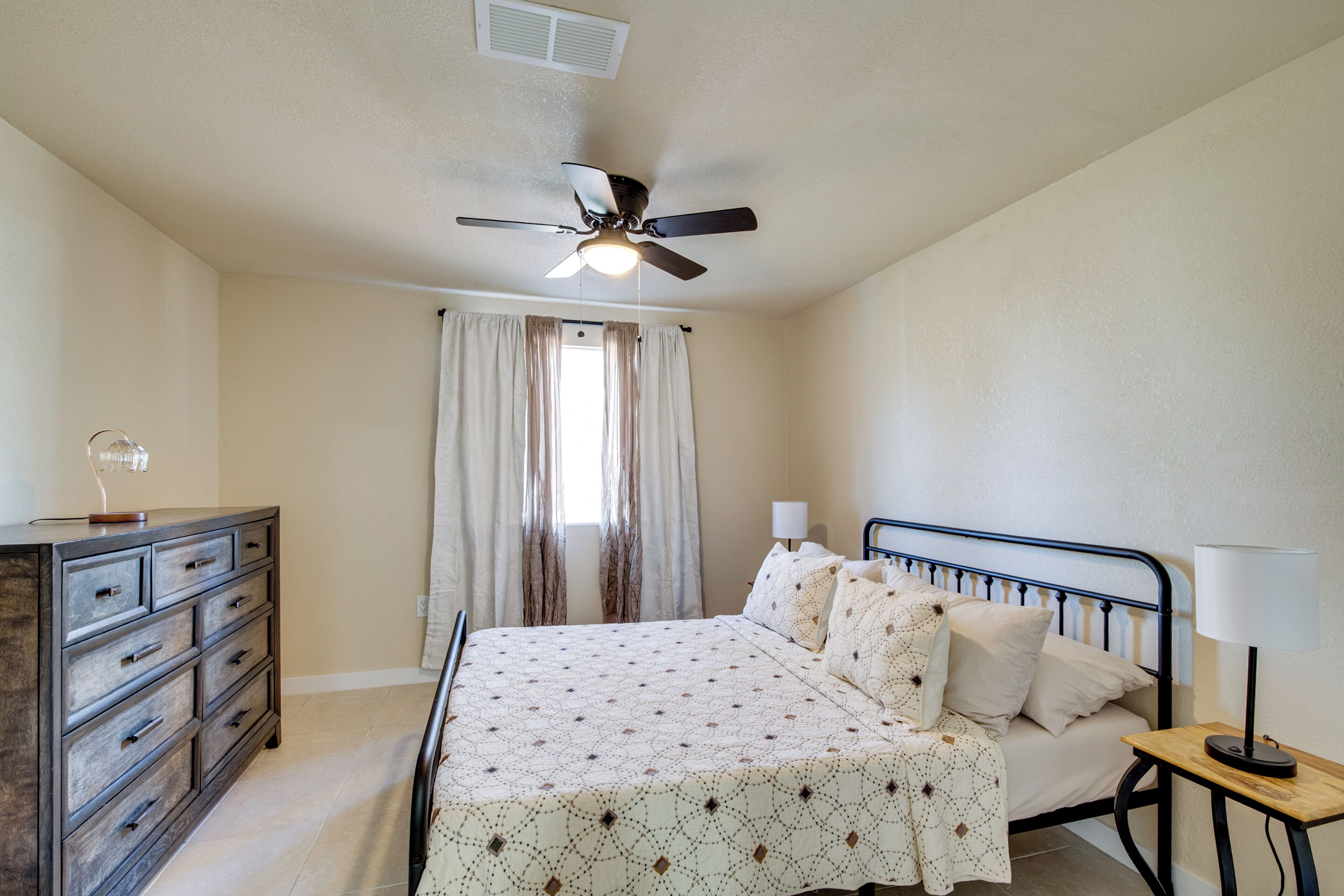 A bedroom with a bed, a dresser, a ceiling fan, and a window with curtains.