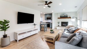 Un salon moderne doté d’un téléviseur à écran plat, d’un ventilateur de plafond, d’un coin repas avec une cuisine en arrière-plan et d’une cheminée.
