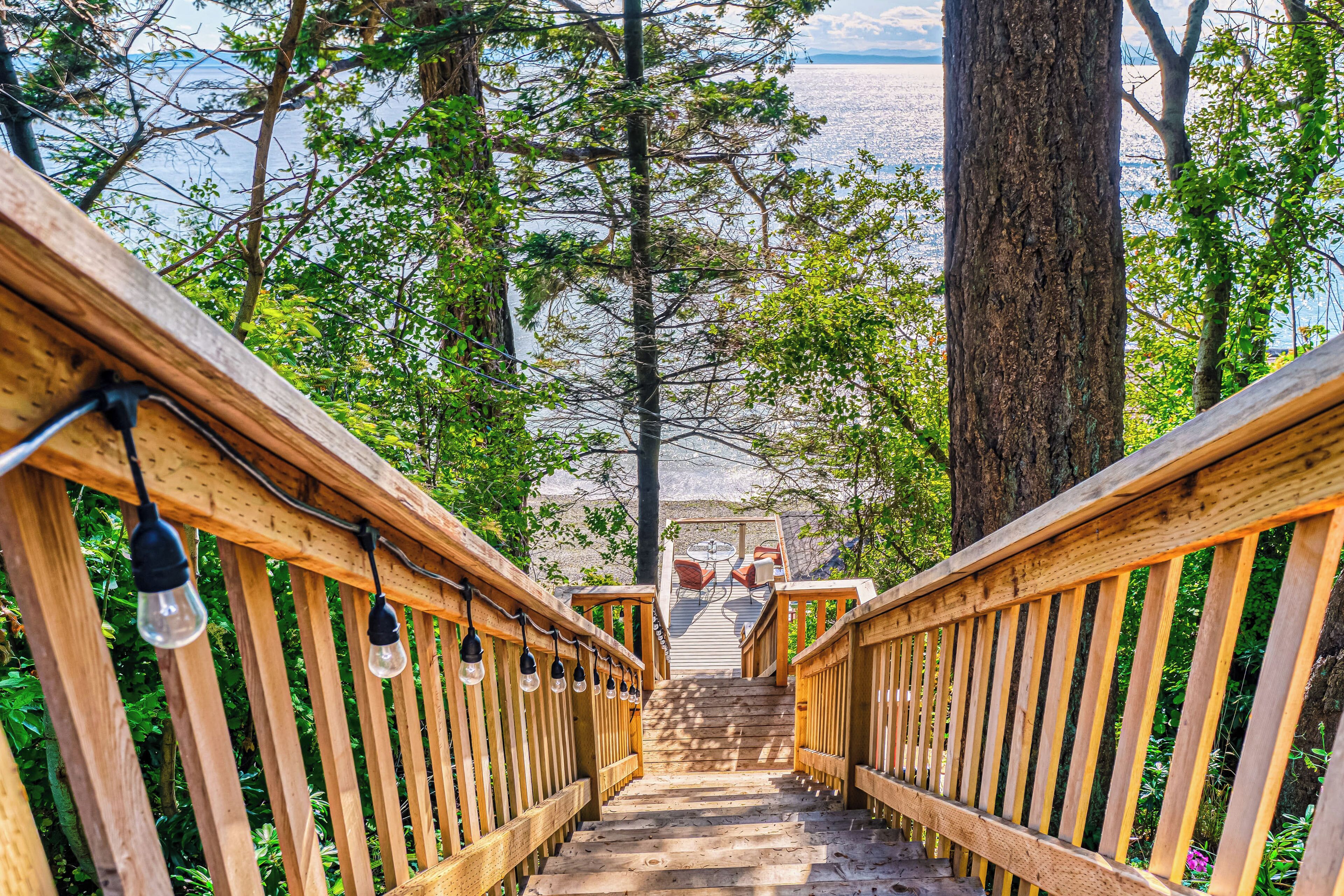 A wooden staircase with railings and hanging light bulbs, leading to a seating area with chairs and a table, surrounded by trees and a view of a body of water.