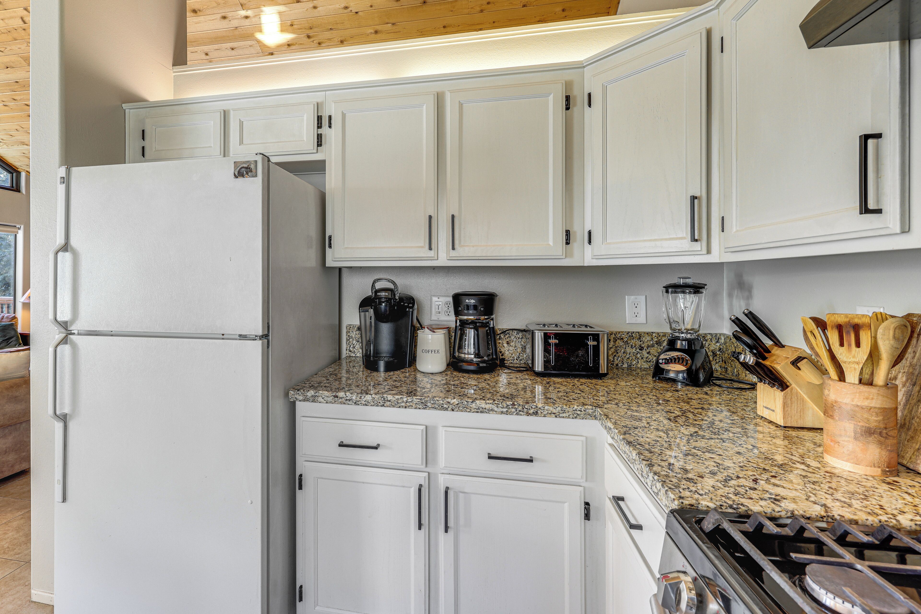 Una cocina con armarios blancos, encimera de granito, refrigerador, cafetera, tostadora, licuadora y estufa.