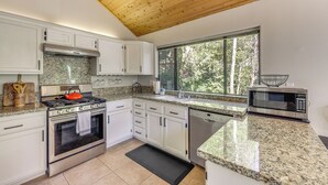Une cuisine moderne avec des armoires blanches, des appareils en acier inoxydable, des comptoirs en granit et une grande fenêtre offrant une vue sur les arbres.