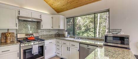 A modern kitchen with white cabinets, stainless steel appliances, granite countertops, and a large window providing a view of trees.