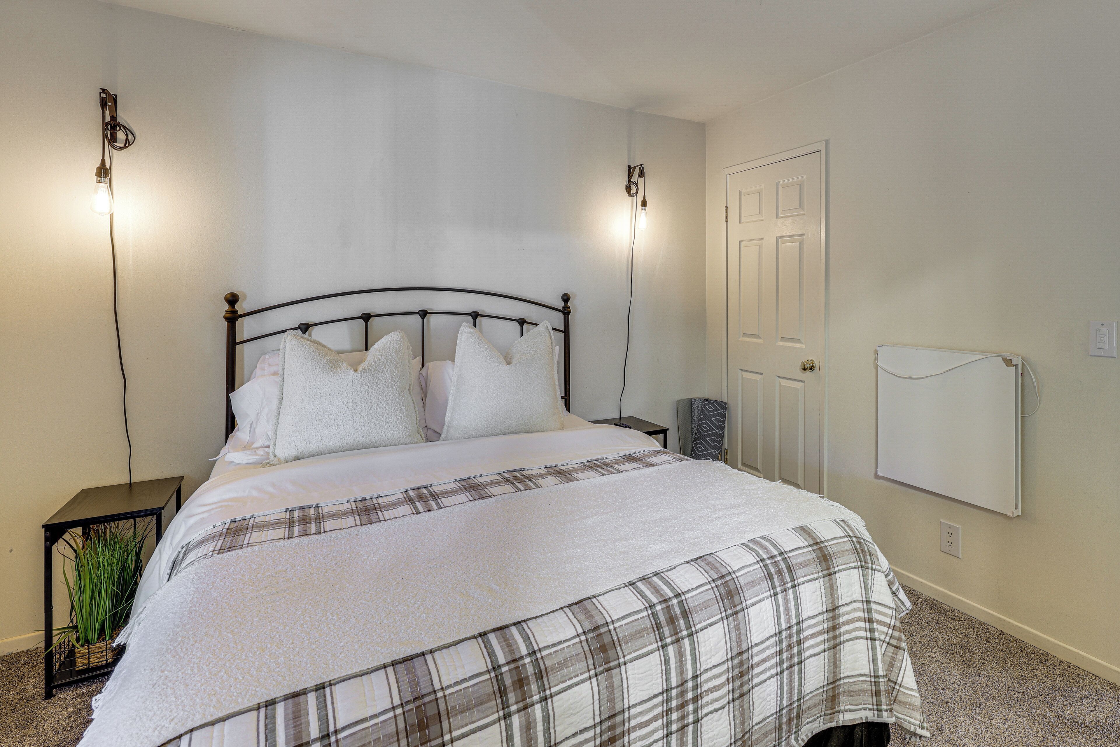 A neatly made bed with a plaid bedspread, two bedside lamps, a small nightstand with a plant, and a white door.