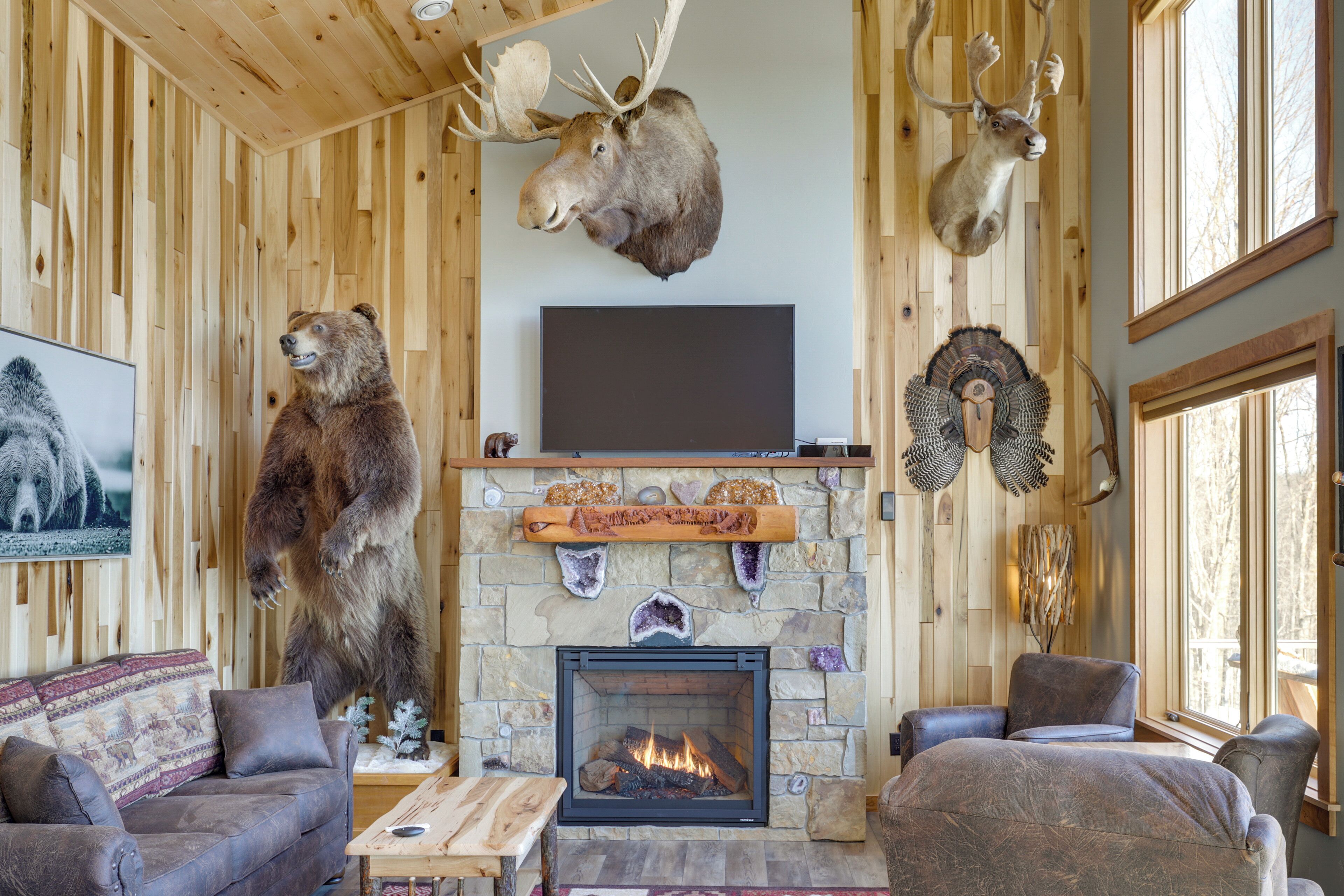 A cozy cabin interior with a stone fireplace, mounted animal heads, and a large flat-screen TV.