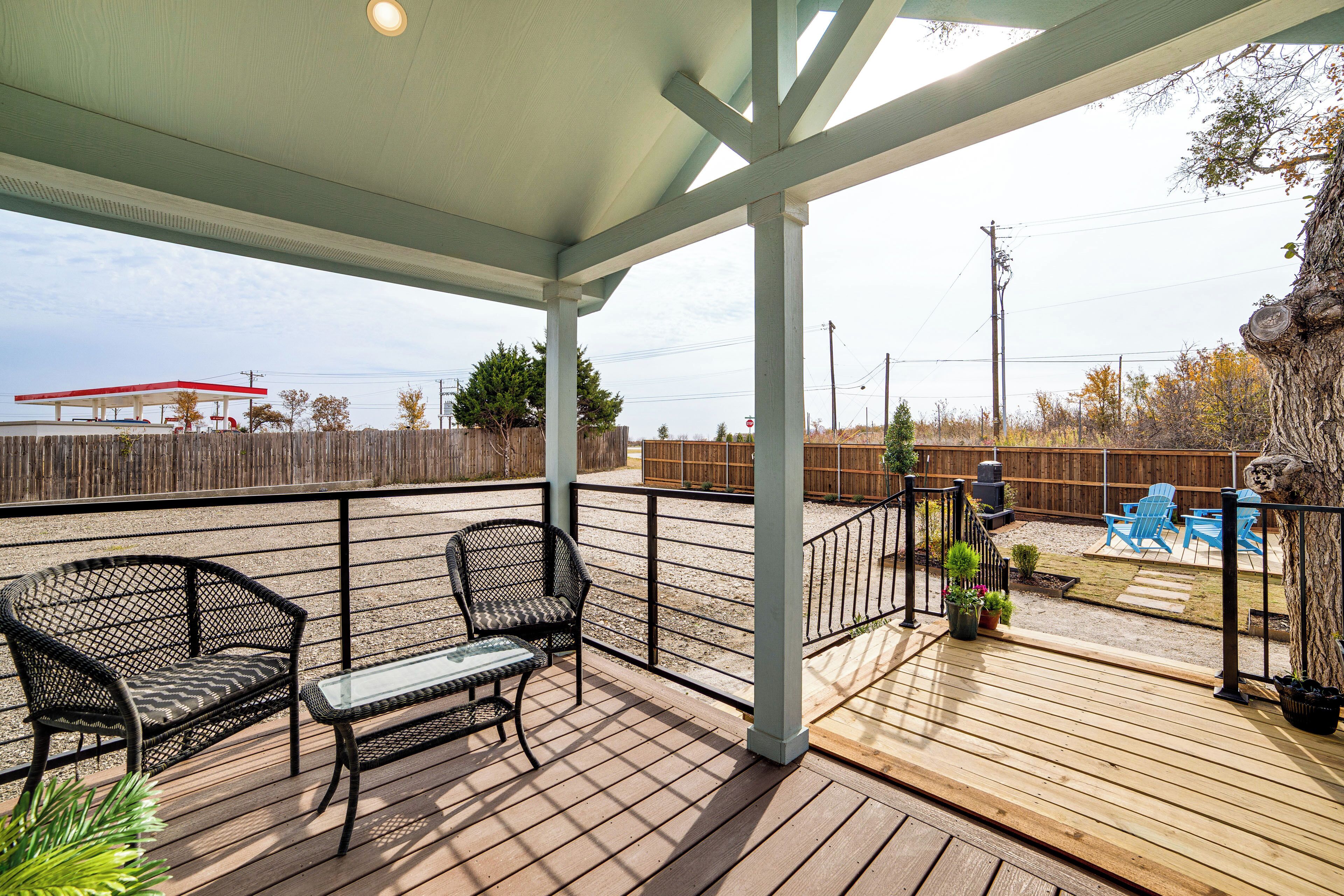 A covered outdoor deck with two chairs and a table, a wooden fence, and a view of a gas station in the background.