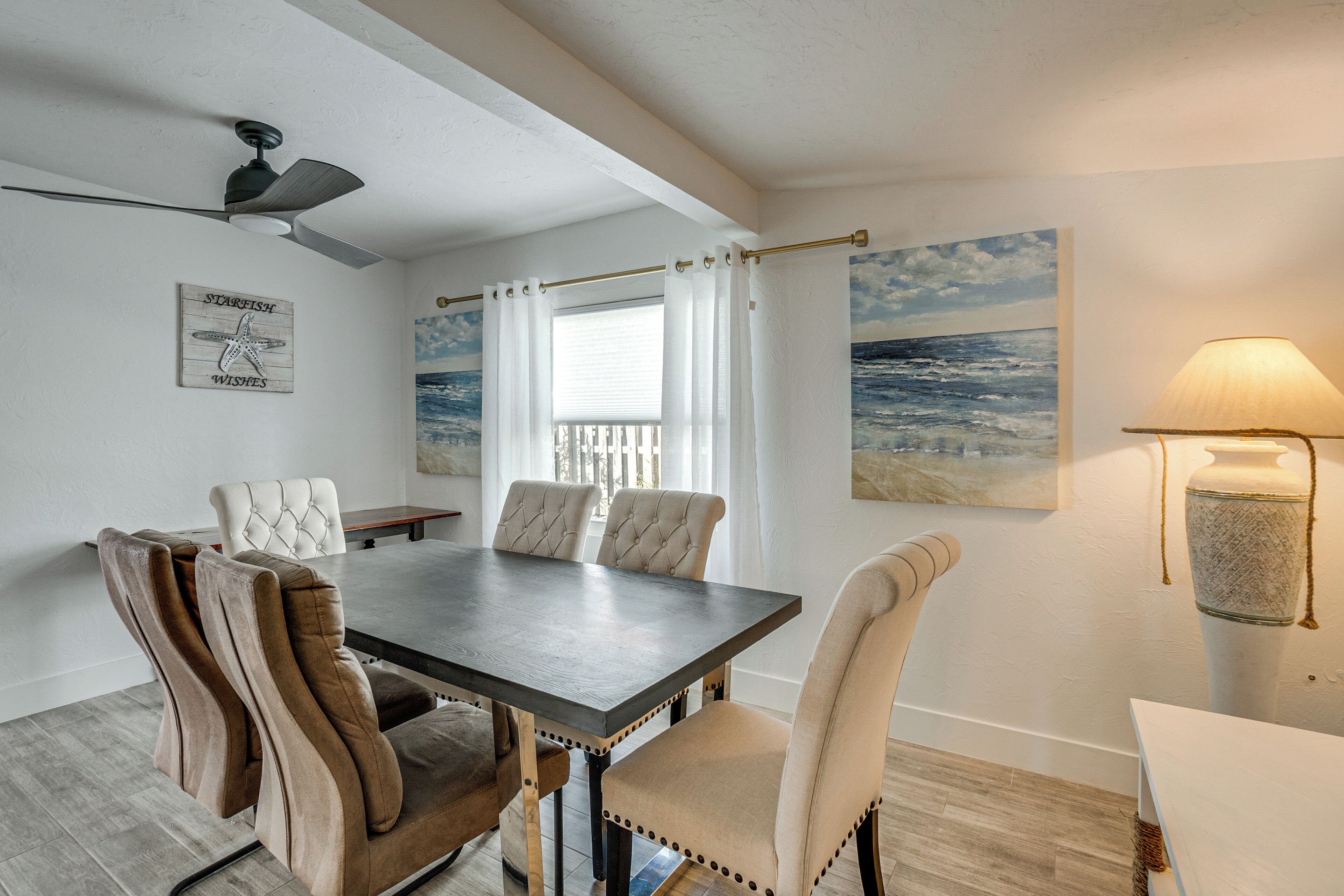 Uma sala de jantar com uma mesa de madeira escura, cadeiras acolchoadas, um ventilador de teto, vista para o oceano através de uma janela com cortinas brancas, e um quadro do mar na parede.