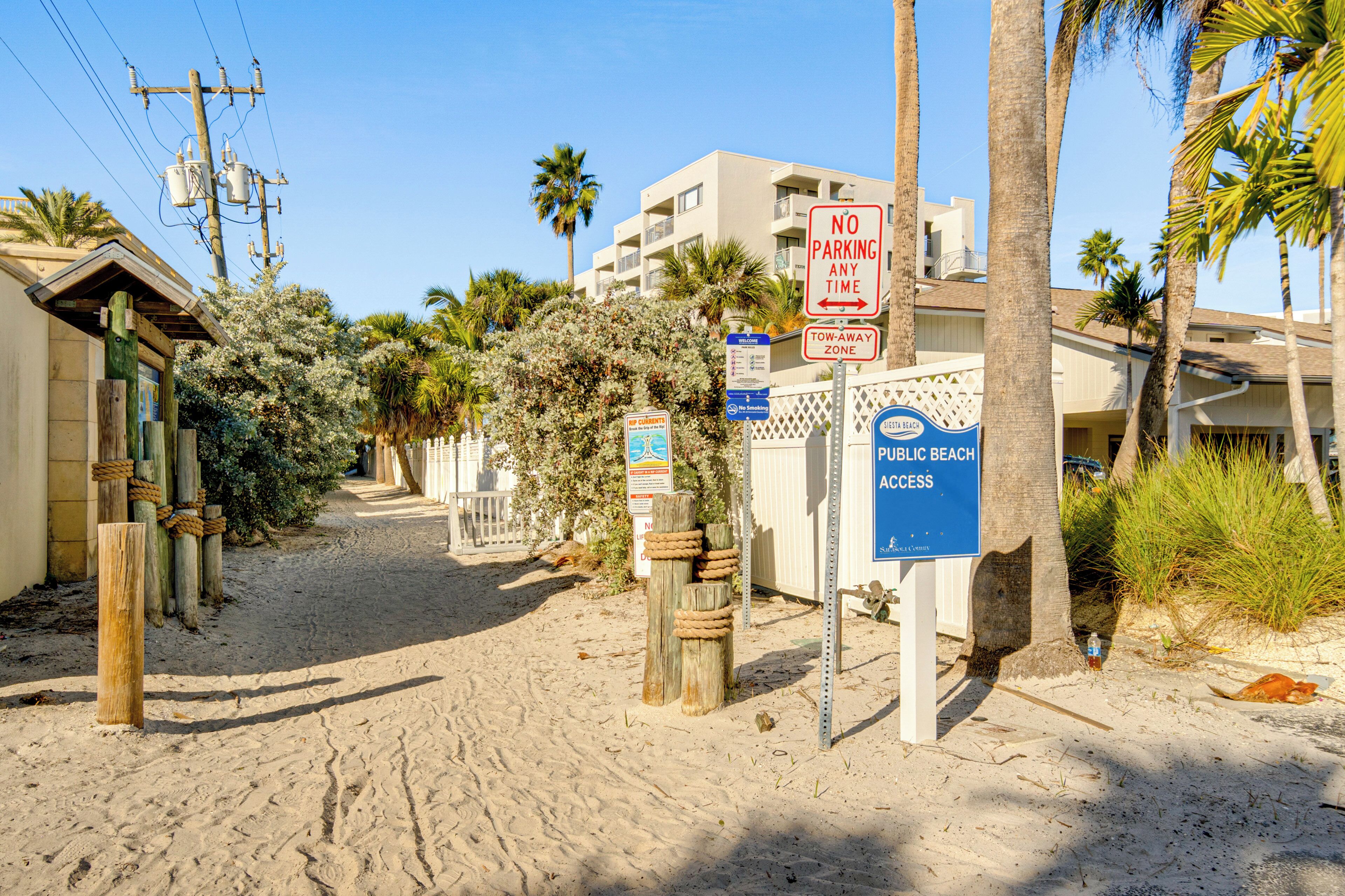 A sandy path leading to a building with a 'No Parking Any Time' sign, a 'Public Beach Access' sign, and a 'Tow-Away Zone' sign.
