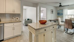 Una cocina con armarios blancos, encimera de granito y un tazón grande. Hay un comedor con mesa y sillas, y vista al océano a través de una ventana.