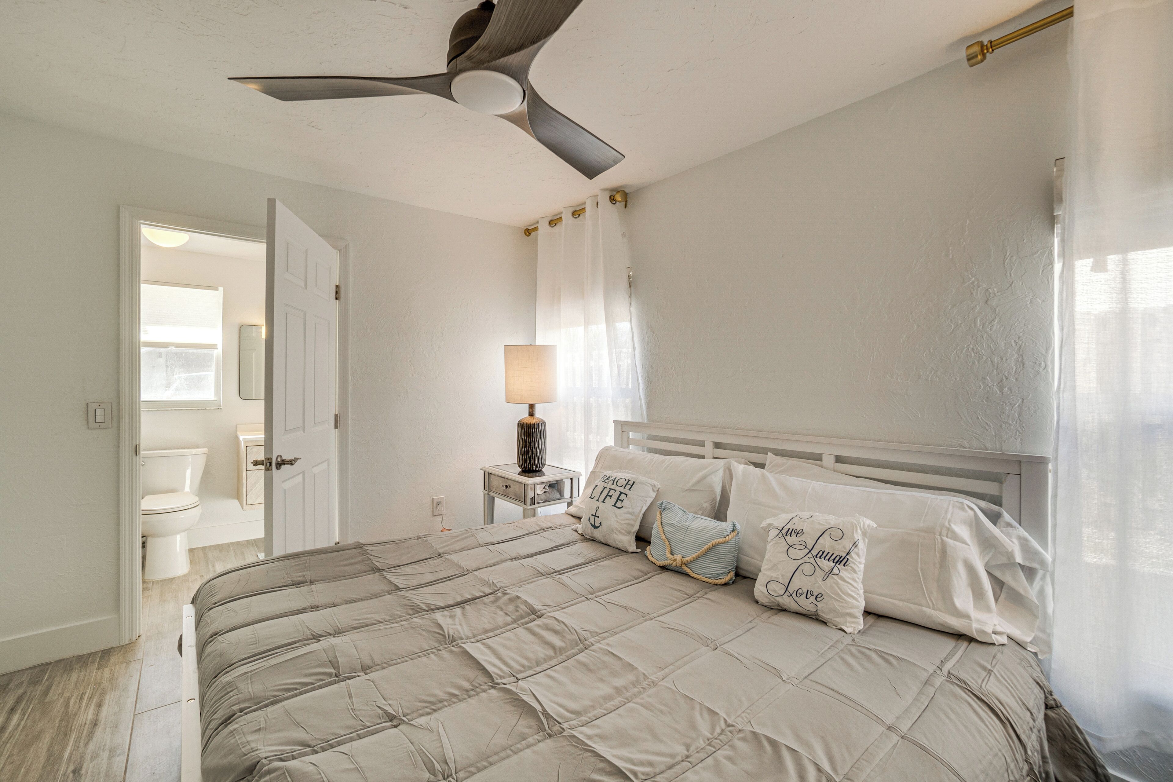 Un dormitorio con cama, ventilador de techo, mesita de noche con lámpara y baño visible a través de una puerta abierta.
