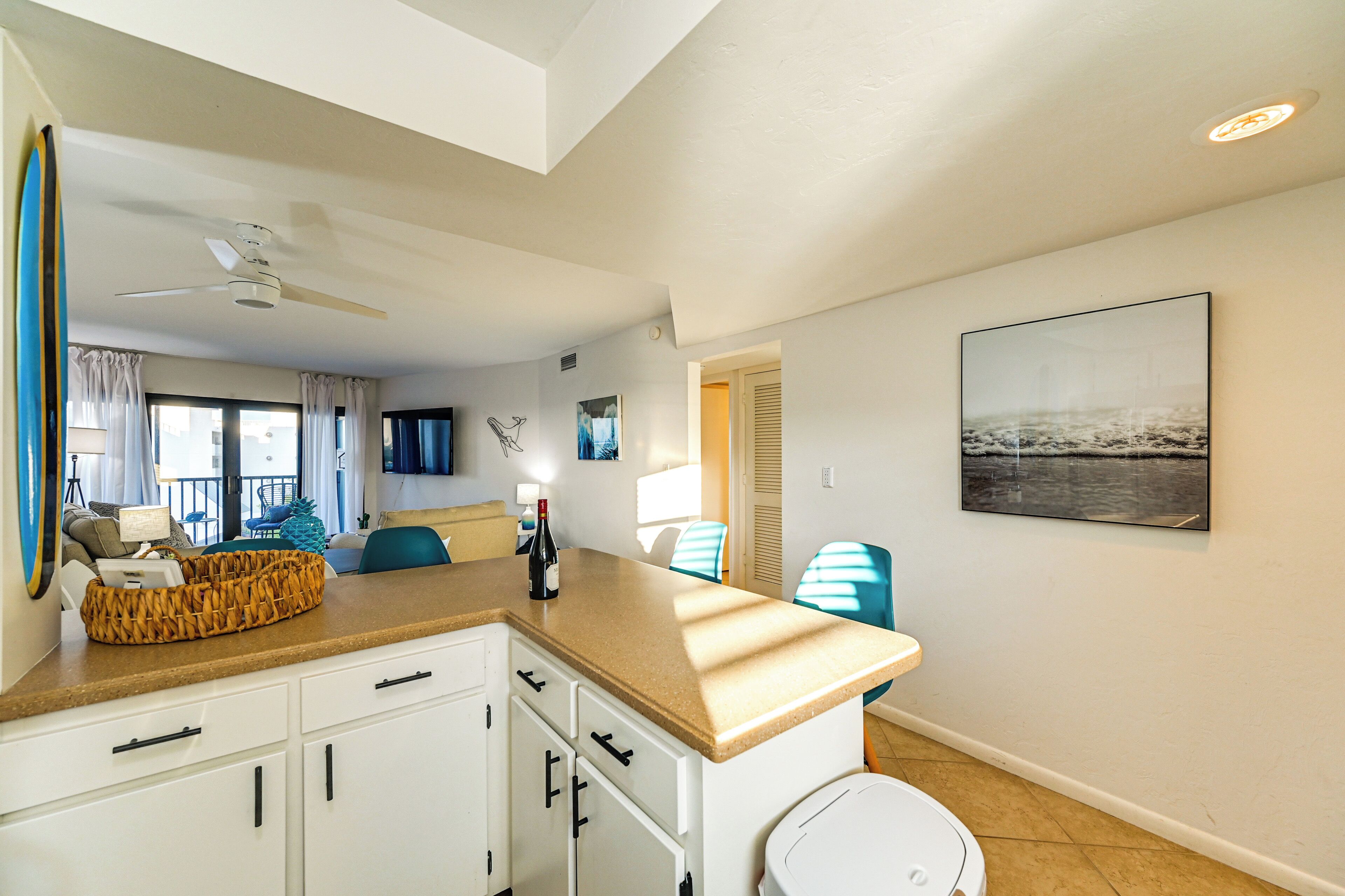 A modern kitchen with white cabinets, a beige countertop, and a ceiling fan. There is a dining area with blue chairs and a table, and a large painting on the wall.
