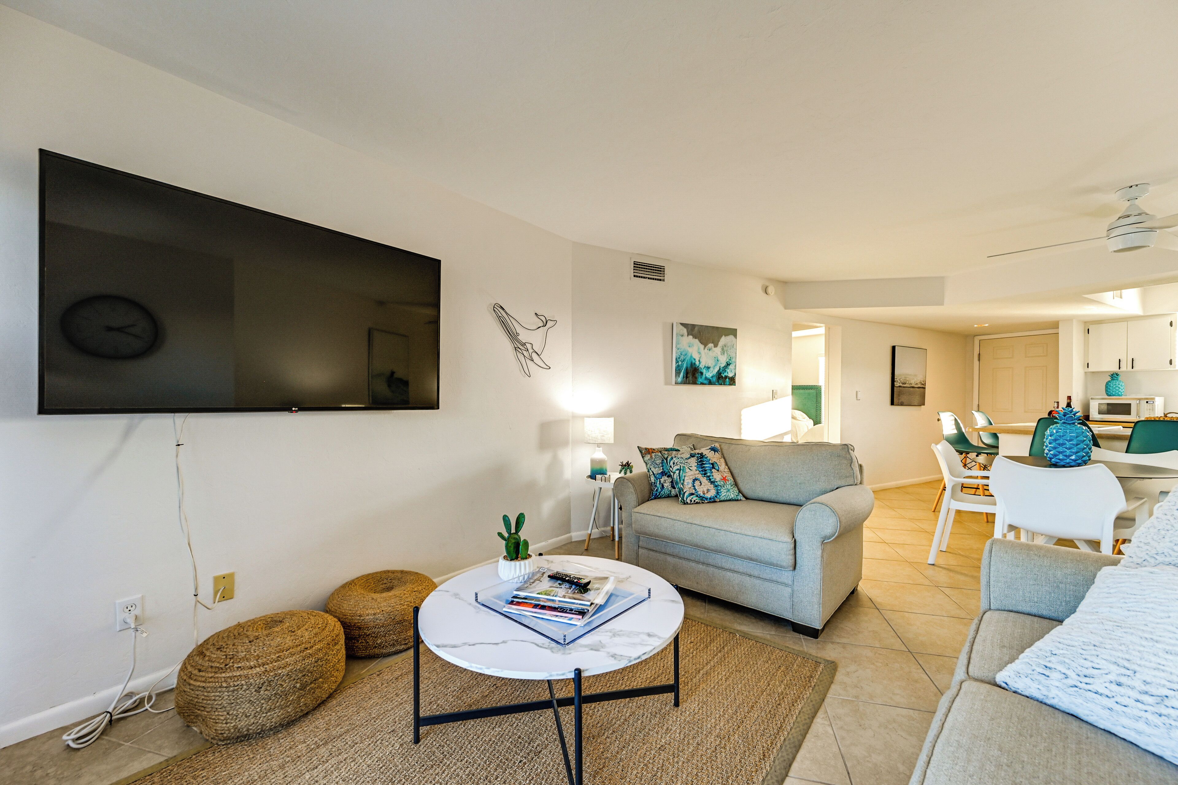 A modern living room with a flat-screen TV, a grey sofa, a coffee table with books, and a dining area in the background.