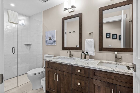 A modern bathroom with a glass shower enclosure, a white toilet, a granite countertop sink, wooden cabinets, and two large mirrors. - Mountains are Calling | Our Penthouse Says Hello (Canmore)