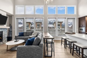 A modern living room with a fireplace, grey sofa, coffee table, and a kitchen area with a bar and stools. - Mountains are Calling | Our Penthouse Says Hello (Canmore)
